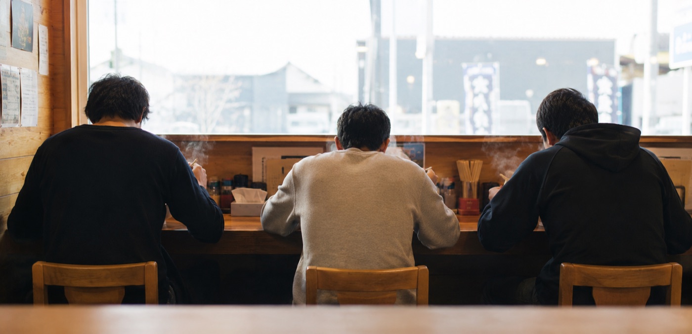 昼、窓に面したカウンターでラーメンを食べるお客さんたちの後ろ姿