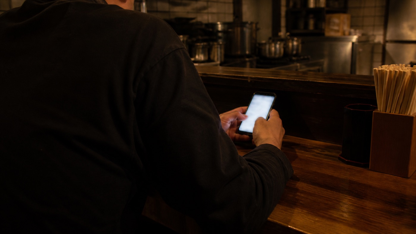 夜の閉店後、カウンターでスマートフォンを見つめる店主の後ろ姿